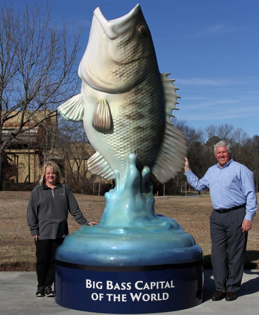 Two people stand beside a large fish statue labeled "Big Bass Capital of the World" in an outdoor setting.