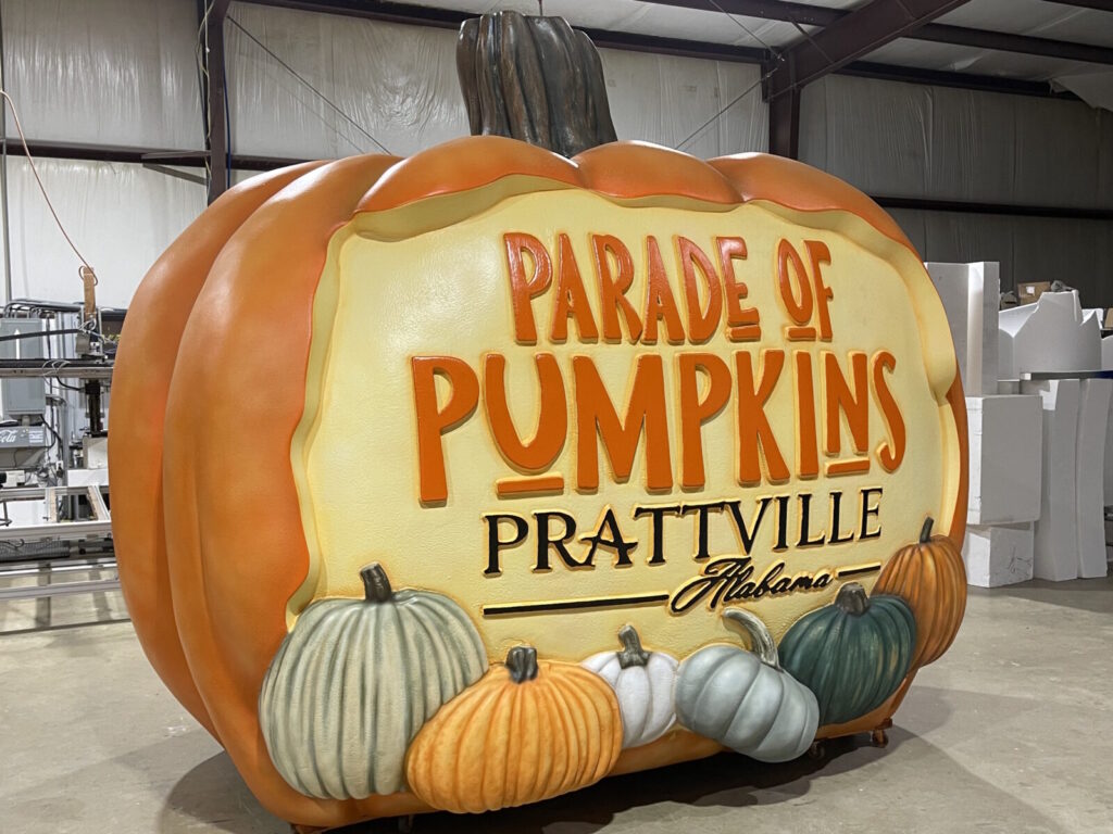 Large pumpkin sculpture with "Parade of Pumpkins Prattville Alabama" text, surrounded by smaller pumpkins, indoors.