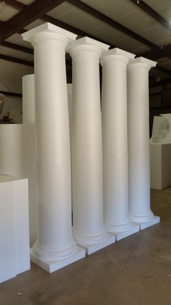 Four large white classical columns stand in a row inside an industrial warehouse space.