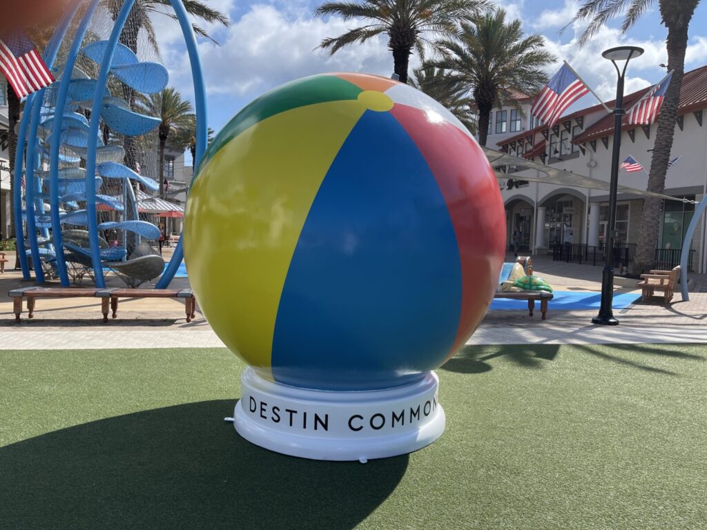 Large colorful beach ball sculpture on a white base labeled "Destin Commons," with palm trees and buildings in the background.