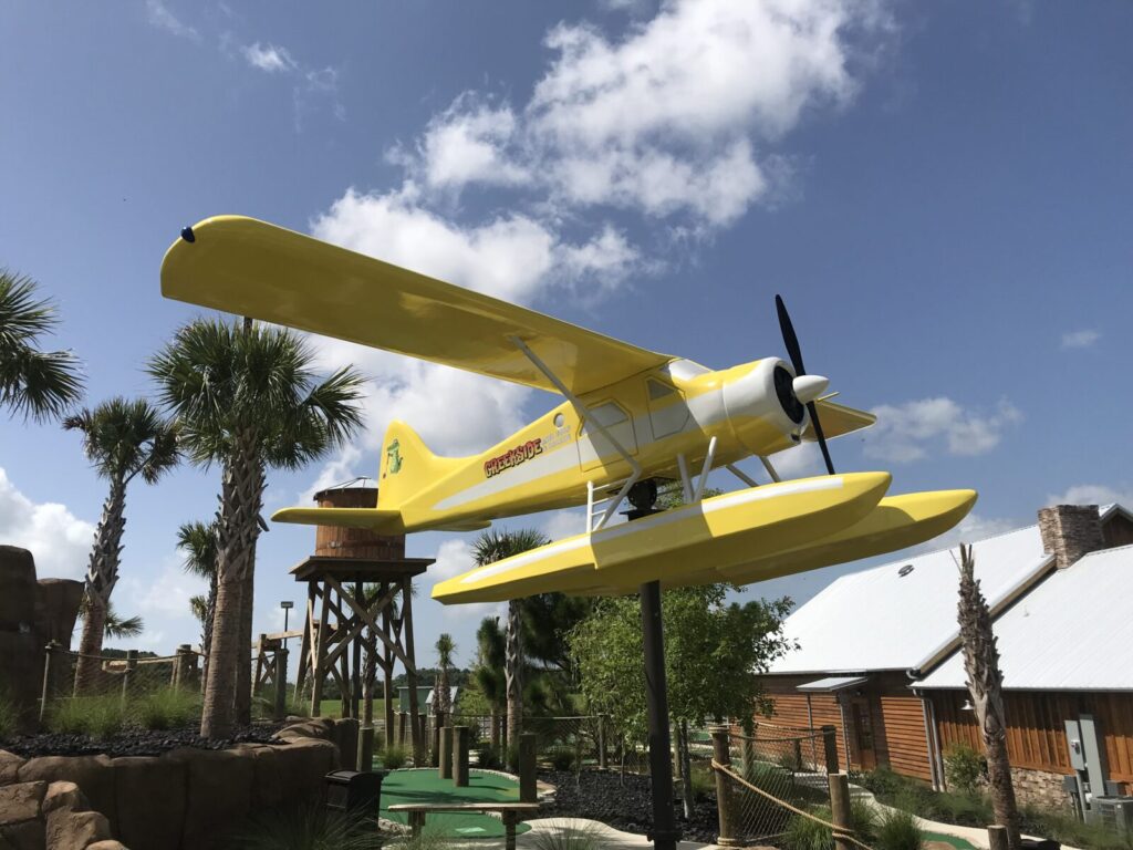 A large yellow seaplane model is displayed on stilts outdoors among palm trees and wooden buildings under a blue sky.