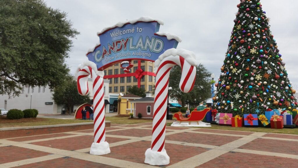 Large "Candyland" sign with giant candy canes stands in plaza near decorated Christmas tree and wrapped gifts.