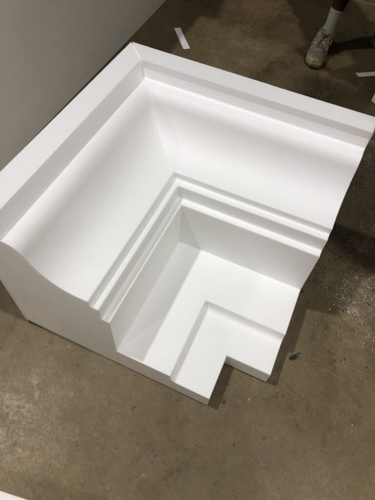 A large, white, L-shaped architectural molding piece sits on a bare concrete floor next to someone's feet.
