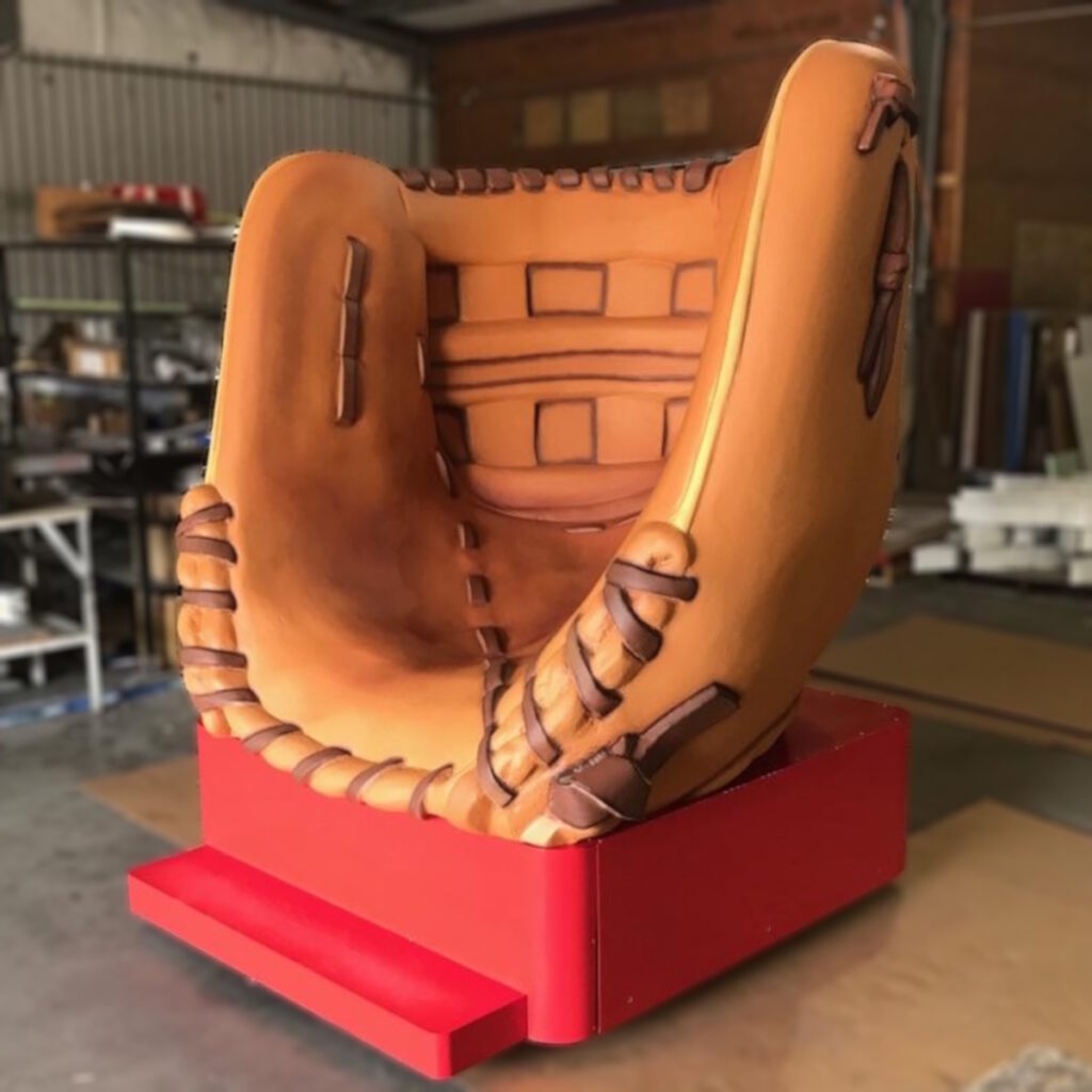 Oversized chair designed to look like a brown baseball glove, set on a red platform inside a warehouse.