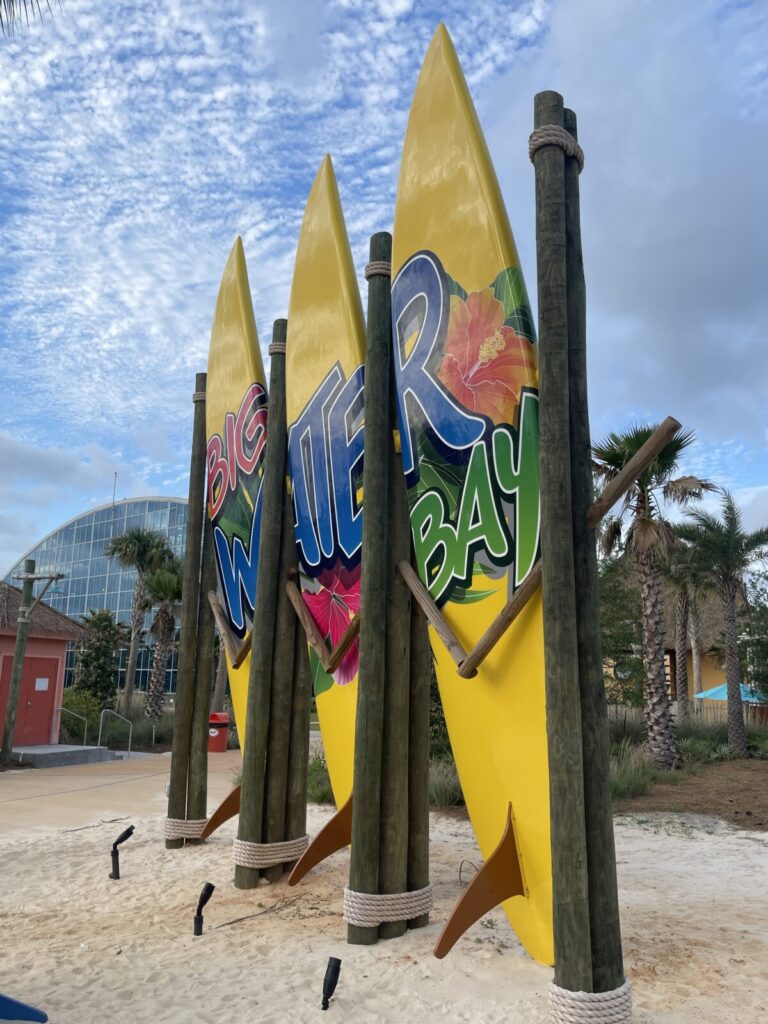 Three large yellow surfboards with the words "BIG WATER BAY" stand upright on a sandy area with palm trees and buildings nearby.