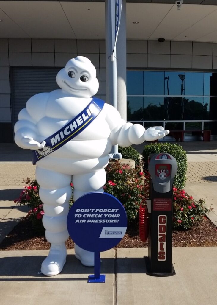 Michelin Man mascot stands beside a tire inflator machine, holding a sign about checking air pressure.