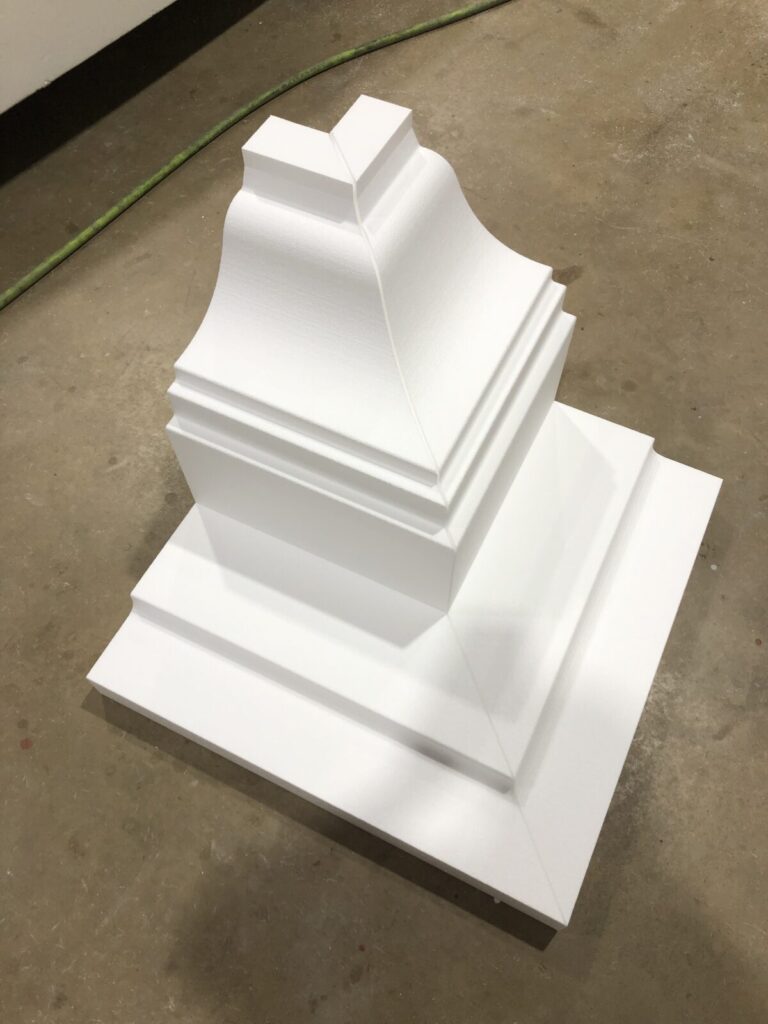 A white, tiered architectural model with clean lines sits on a concrete floor near a green hose.