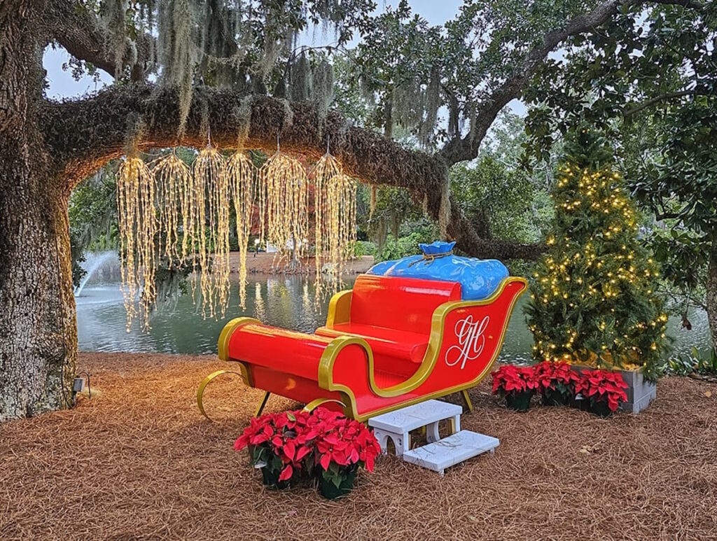 A red sleigh and Christmas tree with lights are set by a pond under a tree with hanging string lights and poinsettias.